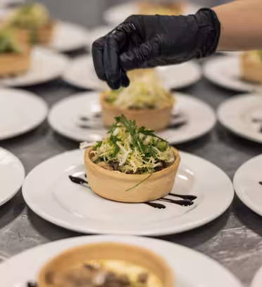 Nærbilde av en kokkes hånd iført en svart hanske, pyntet flere tallerkener med velsmakende terter fylt med grønt og en skvett mørk saus på hvite tallerkener, pent arrangert på en overflate i rustfritt stål.