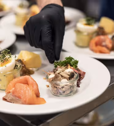 En hånd i en svart hanske plater en gourmetrett med røkt laks, en krukke med blandede grønnsaker, kokt potet og pyntede egg, på en hvit tallerken. Flere plater med lignende arrangement er synlige i bakgrunnen.