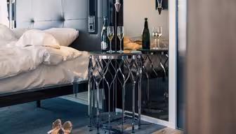 Et moderne soverom med en pent oppredd seng, et elegant sidebord med en vinflaske, to fylte glass og et par høye hæler på gulvet. Innstillingen antyder en sofistikert og intim atmosfære.