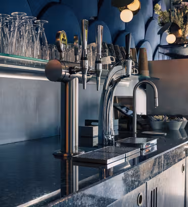 En moderne kafébardisk med granittoverflate har to elegante ølkraner i krom, en rad med omvendte glass på en hylle og en kaffemaskin mot en strukturert blå vegg. Mykt lys gir en varm atmosfære.
