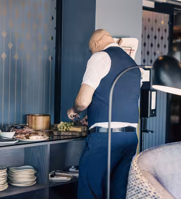 En mann i blå vest med hvit skjorte står ved et buffébord og skjærer mat med en kniv. Bordet rommer tallerkener, boller og en rekke snacks. En lampe står i forgrunnen, og rommet har blågrå vegger med geometriske mønstre.