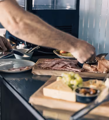 En person i blått forkle serverer pålegg og ost på en tallerken. Bordet viser en treplate med assortert kjøtt, oster, druer og et lite fat med oliven. Plater er stablet i nærheten, og en metallbakgrunn er synlig.