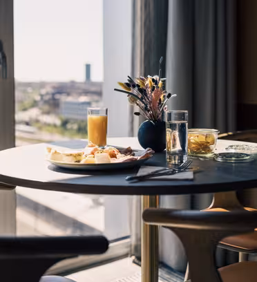 Rundt bord med en tallerken med frokostvarer, inkludert brød og skinke, et glass appelsinjuice og et annet glass med sitronskiver. En liten vase med blomster pynter på bordet. Stort vindu gir utsikt over byen i bakgrunnen.
