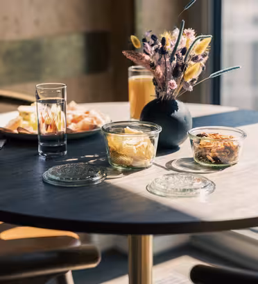 Et solbelyst frokostbord med en vase med tørkede blomster, glass med appelsinjuice og vann, tallerkener med mat og to små krukker med lokk, en med smørbar substans og en annen med nøtter. En serviett og bestikk er plassert i nærheten.