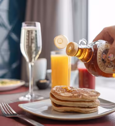 En hånd heller sirup over en bunke pannekaker på en hvit tallerken. Et glass appelsinjuice, en kopp kaffe og en champagnefløyte står på bordet. En uskarp utsikt over byen er synlig gjennom et vindu.
