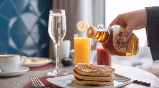 En hånd heller sirup over en bunke pannekaker på en hvit tallerken. Et glass appelsinjuice, en kopp kaffe og en champagnefløyte står på bordet. En uskarp utsikt over byen er synlig gjennom et vindu.