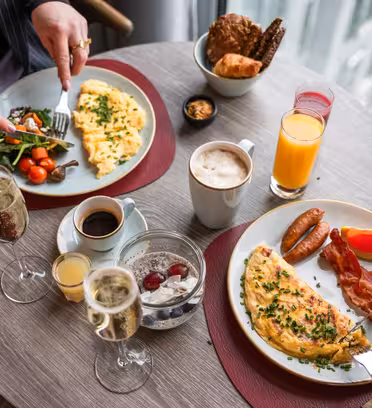 To personer spiser ved et rundt bord. Bordet har tallerkener med omeletter, pølser, bacon, salat og cherrytomater. Det er drinker inkludert juice, kaffe og musserende vin. En brødkurv står også på bordet.
