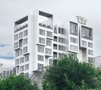 Moderne hvit og grå fleretasjes bygning med takområde, omgitt av grønne trær og mot en overskyet himmel. Strukturen har geometriske design med mange vinduer og et takskilt.