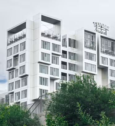 Moderne hvit og grå fleretasjes bygning med takområde, omgitt av grønne trær og mot en overskyet himmel. Strukturen har geometriske design med mange vinduer og et takskilt.