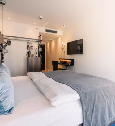 Moderne hotellrom med en pent oppredd dobbeltseng, grått teppe og blå aksentpute. En veggmontert TV vender mot sengen. Det er et lite skrivebord med en stol, en tregarderobe og en kaffemaskin til venstre. Myk belysning skaper en koselig atmosfære.