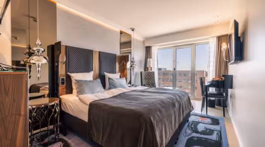 Et moderne hotellrom med king-size-seng, grått sengetøy og to speil bak sengegavlen. Rommet har veggmontert TV, skrivebord med stol, gulv-til-tak-vinduer og stilige pendellamper.