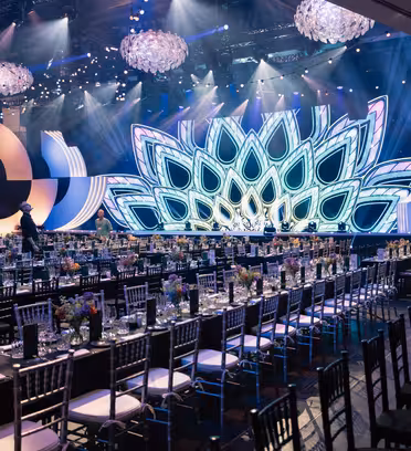 En stor, utsmykket arrangementssal med elegante, opplyste dekorasjoner og et påfuglfjærdesign på scenen. Rader med bord er dekket med stoler, blomsterdekor og servise. Overhead, glitrende lysekroner legger til stemningen.