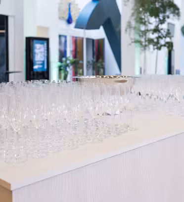 Et stort antall tomme champagneglass er ordnet pent på et hvitt bord. Bakgrunnen har grøntområder og moderne innredning i et romslig, godt opplyst interiør.