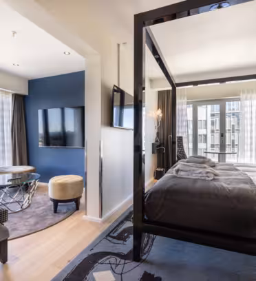Et moderne hotellrom med himmelseng med hvitt og grått sengetøy, sittegruppe med to stoler og salongbord med glassplate, veggmontert TV og store vinduer med utsikt over byen og rene gardiner.