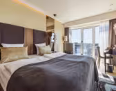 Moderne hotellrom med 2 enkeltsenger, brun og gullinnredning, og et stort vindu med utsikt over byen. Et skrivebord med stol er plassert nær vinduet, og en hengende lampe gir et dekorativt preg. Naturlig lys fyller rommet.