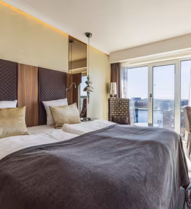 Moderne hotellrom med 2 enkeltsenger, brun og gullinnredning, og et stort vindu med utsikt over byen. Et skrivebord med stol er plassert nær vinduet, og en hengende lampe gir et dekorativt preg. Naturlig lys fyller rommet.