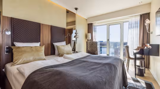 Moderne hotellrom med 2 enkeltsenger, brun og gullinnredning, og et stort vindu med utsikt over byen. Et skrivebord med stol er plassert nær vinduet, og en hengende lampe gir et dekorativt preg. Naturlig lys fyller rommet.