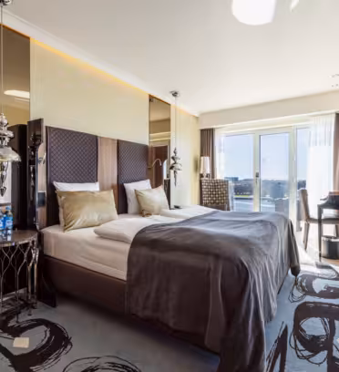 Elegant hotellrom med moderne design. Har en stor seng med beige og grått sengetøy, hodegavl med speil, dekorative sidebord, arbeidsplass nær vinduet og utsikt over byen. Naturlig lys oversvømmer rommet gjennom glassdører som fører til en balkong.