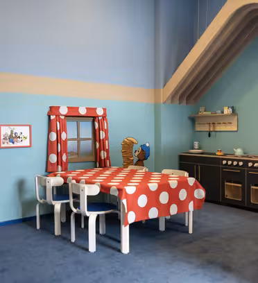 Et lekerom for barn har tegneserie-temadesign med blått og blågrønt fargevalg. Rommet inkluderer et lekekjøkken med svarte skap, et bord med en rød og hvit prikkete duk og tegneseriedekor på veggene.