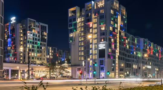Nattutsikt over en moderne hotellbygning med et skilt med "Tivoli". Vinduene er opplyst i ulike farger. Gatelys og trafikklys er synlige, med uskarpe billys som indikerer bevegelse på veien.