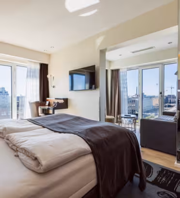 Et moderne hotellrom med stor seng, puter og et grått teppe. Det er en veggmontert TV, et lite skrivebord med stol og en sittegruppe ved vinduet med utsikt over bygninger. Gulv-til-tak-vinduer slipper inn naturlig lys.