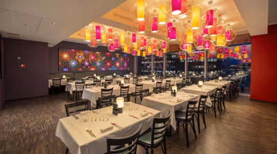 Et moderne restaurantinteriør med pent ordnede bord dekket av hvite duker. Fargerike lykter henger fra taket. Et levende veggmaleri dekorerer den ene veggen, og store vinduer viser et nattlig bybilde.