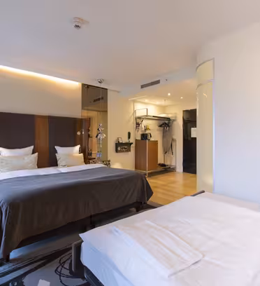 Et moderne hotellrom med to pent oppredde senger, med hvitt sengetøy og mørkegrå detaljer. Rommet har tregulv, veggmontert TV og moderne belysning. Et stort speil og minimalistisk innredning skaper en romslig og ren atmosfære.