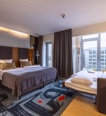 Et moderne hotellrom med to senger, elegante møbler, store vinduer med utsikt over byen, mørke gardiner, et mønstret teppe og stemningsbelysning. Et skrivebord og en stol er plassert i nærheten av vinduet.