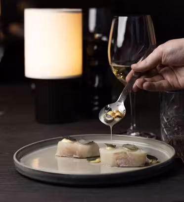 En hånd som holder en skje drypper saus over to fiskestykker pyntet med skiver på en tallerken. Vinglass og en lysende lampe skaper en elegant spisestue på et mørkt bord.
