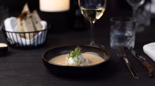 En bolle med kremet suppe pyntet med urter står på et mørkt bord. Ved siden av er et glass hvitvin, et vannglass, en gaffel og en kniv. I bakgrunnen er det en kurv med brød og et svakt tent lys.