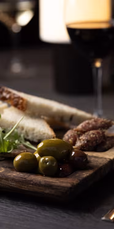 En treplate med assortert charcuterie: oppskåret kjøtt, oliven, ost og brød. Friske grønnsaker pynter fatet. I nærheten står et glass rødvin og et krystallglass på det mørke trebordet, dekket med kniv og gaffel og en sammenrullet serviett.
