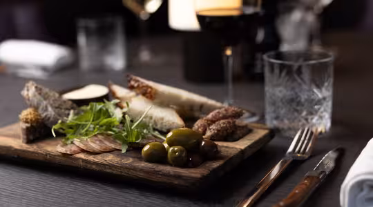 En treplate med assortert charcuterie: oppskåret kjøtt, oliven, ost og brød. Friske grønnsaker pynter fatet. I nærheten står et glass rødvin og et krystallglass på det mørke trebordet, dekket med kniv og gaffel og en sammenrullet serviett.