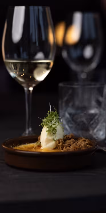 Et svakt opplyst bord med en dessert i en bolle pyntet med en frisk urt. Ved siden av står et vinglass med hvitvin, et glass isvann, bestikk, en pepperkvern, en rullet serviett og en brennende lampe.