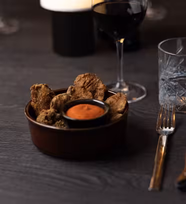 En bolle med stekt sopp med dipsaus på et mørkt bord, ledsaget av et glass rødvin og et glass vann. En gaffel og kniv er plassert ved siden av bollen. Innstillingen er svakt opplyst, noe som antyder en koselig spiseatmosfære.