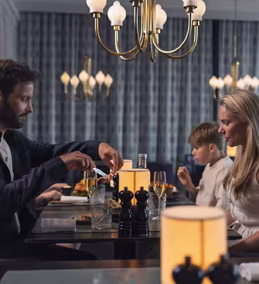 En familie spiser på en restaurant. En mann bruker en pepperkvern, og en kvinne og et barn sitter ved bordet. Elegante lysekroner og bordlamper gir dempet belysning i moderne, stilige omgivelser.