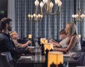 En familie på fire sitter ved et restaurantmiddagsbord. Foreldrene står overfor hverandre, med to barn som sitter mellom dem. De er engasjert i samtale og er omgitt av elegante lysekroner og varm belysning.