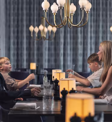 En familie på fire sitter ved et restaurantmiddagsbord. Foreldrene står overfor hverandre, med to barn som sitter mellom dem. De er engasjert i samtale og er omgitt av elegante lysekroner og varm belysning.