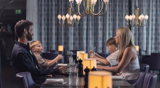 En familie på fire sitter ved et restaurantmiddagsbord. Foreldrene står overfor hverandre, med to barn som sitter mellom dem. De er engasjert i samtale og er omgitt av elegante lysekroner og varm belysning.