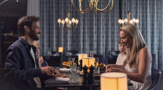 En mann og kvinne spiser sammen på en elegant restaurant med barn bak seg. De smiler og nyter et måltid, opplyst av varm lysekronebelysning.