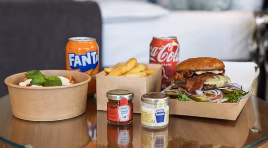 Et glassbord med et måltid inkludert en burger med salat og bacon, pommes frites i en boks, en pastarett med basilikum og krydder. To bokser med brus, Fanta og Coca-Cola, står i bakgrunnen, med en sløret sofa bak seg.