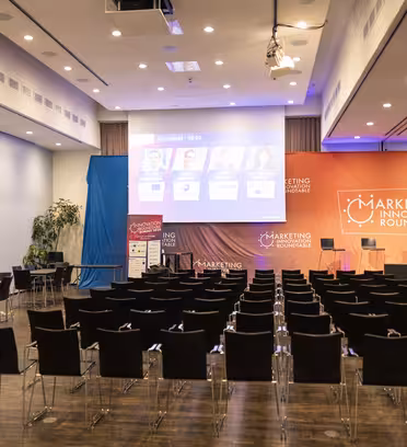 Et tomt konferanserom satt opp med rader med svarte stoler vendt mot en scene. Scenen har et banner med "Markedsføringsinnovasjon Roundtable." Flere skjermer viser høyttalerinformasjon. Rommet er godt opplyst, med kameraer plassert for opptak.