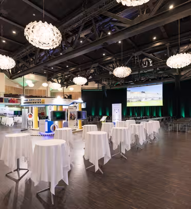 Vidvinkelvisning av et romslig konferanserom med elegante runde bord drapert i hvite kluter. Store lysekroner henger i taket, og en stor skjerm på scenen viser en presentasjon. Deltakerne er synlige i uformelle antrekk.