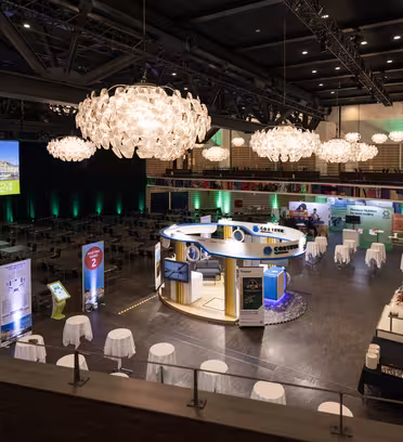 Romslig konferansesal med elegante lysekroner, med en sentral sirkulær messe, omkringliggende utstillinger og mange bord med hvite duker. En stor skjerm viser «Velkommen 2024». Rommet er svakt opplyst, og skaper en innbydende atmosfære.