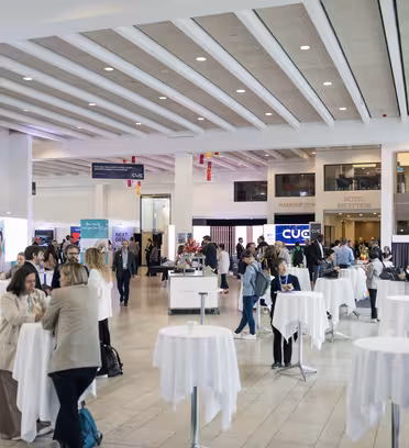 Et sterkt opplyst konferanserom med grupper av mennesker i nettverk rundt hvite runde bord. Bannere og skilt er synlige i bakgrunnen og rommet har en moderne design med stor takhøyde og naturlig lys.