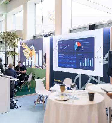 Et sterkt opplyst konferanseoppsett som viser frem digitale skjermer med grafer og diagrammer. Deltakerne samhandler rundt bordene med hvite duker. Potteplanter og et stort vindu gir et naturlig element til det moderne innendørsrommet.