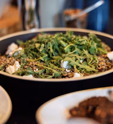 En skål med linsesalat toppet med ruccola og ostebiter. Andre retter, inkludert en tallerken med dip og en dessert med sjokoladebiter, er delvis synlige i forgrunnen og bakgrunnen.