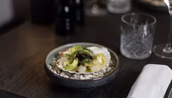 En gourmetrett sitter i en mørk bolle på et restaurantbord. Retten har grønt og urter på toppen av kamskjell på en seng av småstein. I nærheten finner du en brettet hvit serviett, et tomt glass, en pepperkvern og et mykt glødende stearinlys.