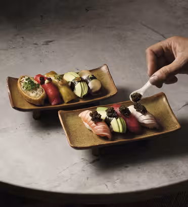 En hånd legger forsiktig kaviar på et stykke sushi på et rektangulært keramisk fat. Begge skålene består av forskjellige fargerike sushibiter arrangert på en strukturert steinoverflate.