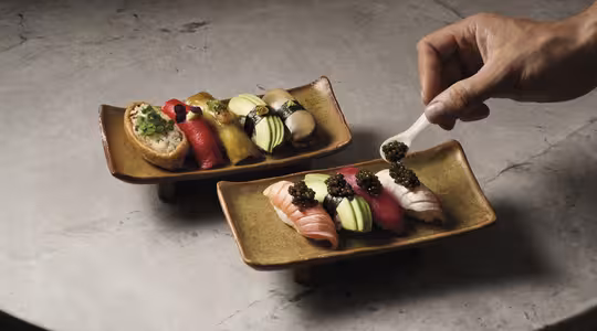 En hånd legger forsiktig kaviar på et stykke sushi på et rektangulært keramisk fat. Begge skålene består av forskjellige fargerike sushibiter arrangert på en strukturert steinoverflate.