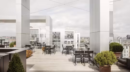 Takterrasse med moderne arkitektur med sølvstrukturer og treterrasse. Den har uteservering med svarte bord og stoler, omgitt av potteplanter. Utsikten inkluderer bybygninger under en overskyet himmel.